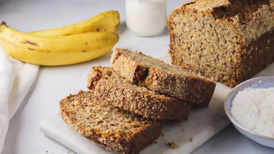 Freshly sliced coconut banana bread on a marble board with bananas.