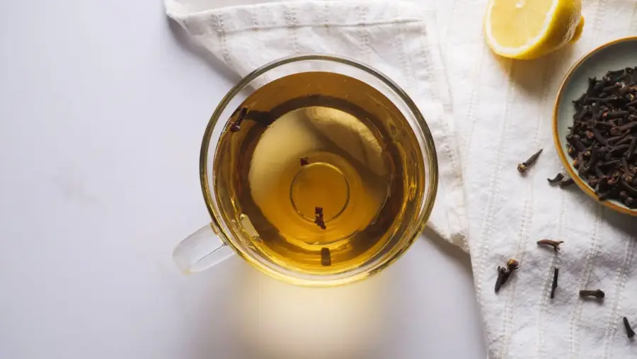Clove tea into a cup with a black tea bag, a lemon slice, a clove, honey, and a teapot nearby.