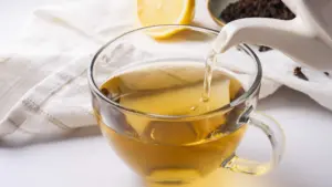 Pouring clove tea into a cup with a lemon slice, clove, and honey nearby.