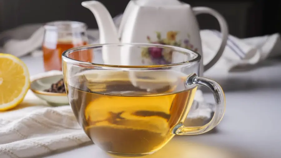 Clove tea into a cup with a black tea bag, a lemon slice, a clove, honey, and a teapot nearby.