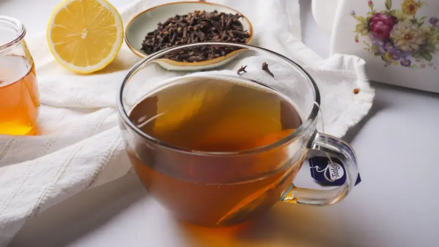 Clove tea into a cup with a black tea bag, a lemon slice, a clove, honey, and a teapot nearby.