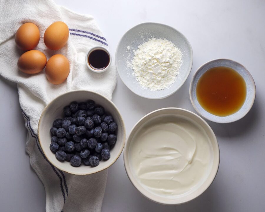 Baking ingredients: eggs, blueberries, yogurt, and more on table.