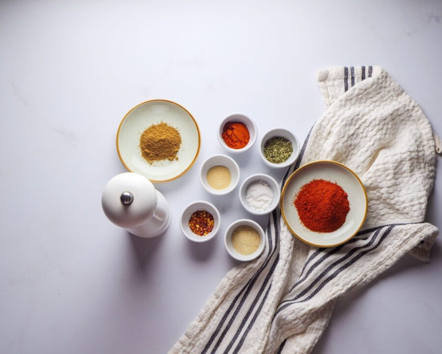 Assorted spices in bowls with a pepper mill on a white cloth.