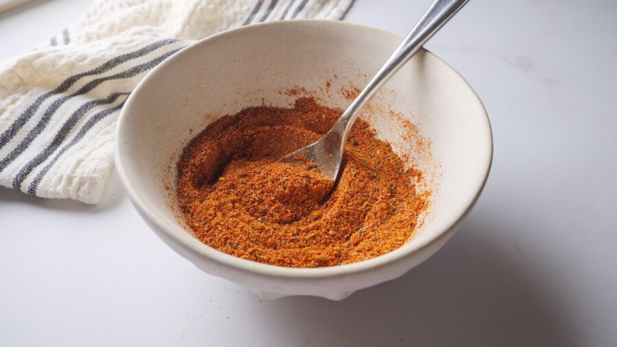 Bowl of mixed taco spices with a spoon on a white surface.