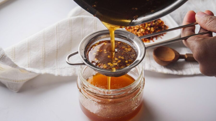 Filtering hot honey sauce through a strainer into a glass jar.