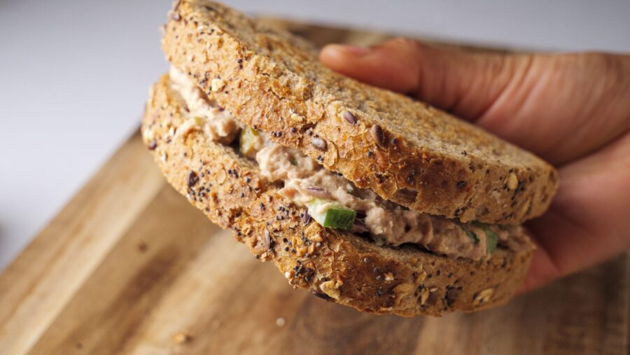 Whole-grain tuna salad sandwich in hand on a wooden board.